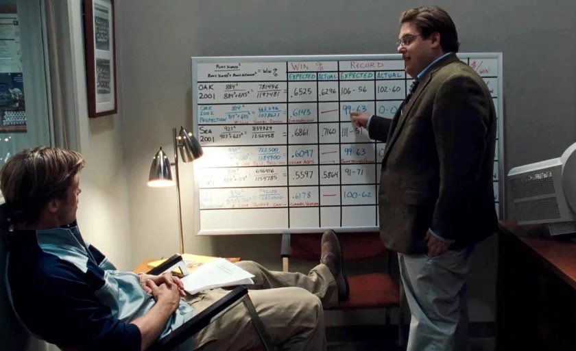 Moneyball whiteboard scene: Peter Brand presenting a data-heavy case to a reclined, sceptical Billy Beane